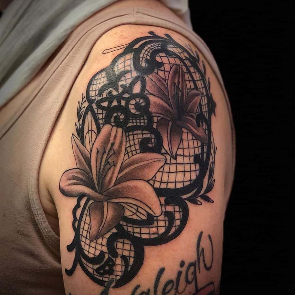 Upper arm tattoo featuring elegant lilies intertwined with intricate black lace patterns and ornamental filigree details.