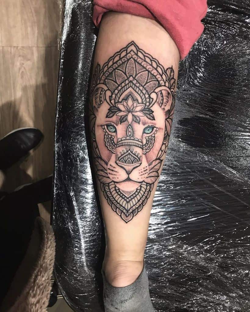 Intricate mandala-style lioness tattoo on the leg, featuring bold black outlines, geometric details, and striking blue eyes for a powerful look.
