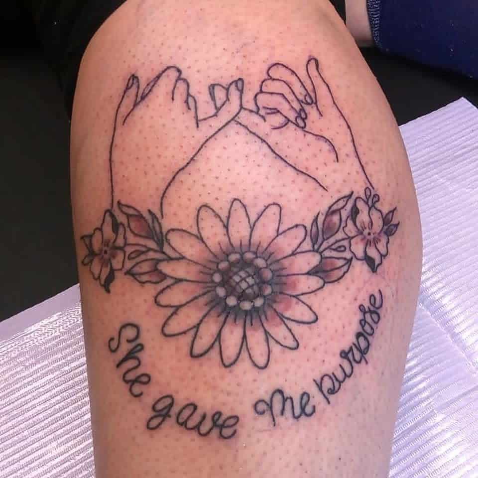 A tattoo of two intertwined hands above a sunflower and small flowers with the text "She gave me purpose" beneath