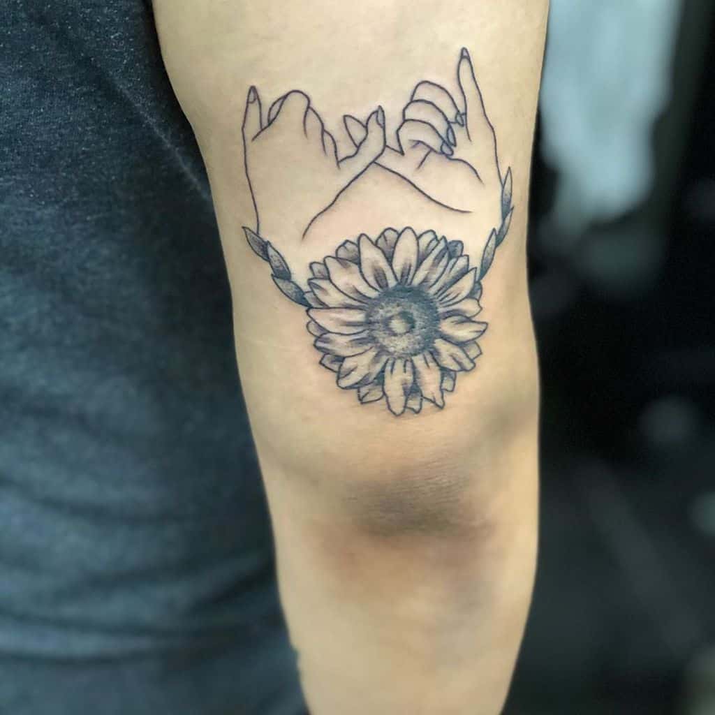 Line tattoo of two hands making a pinky promise above a sunflower, located on an upper arm