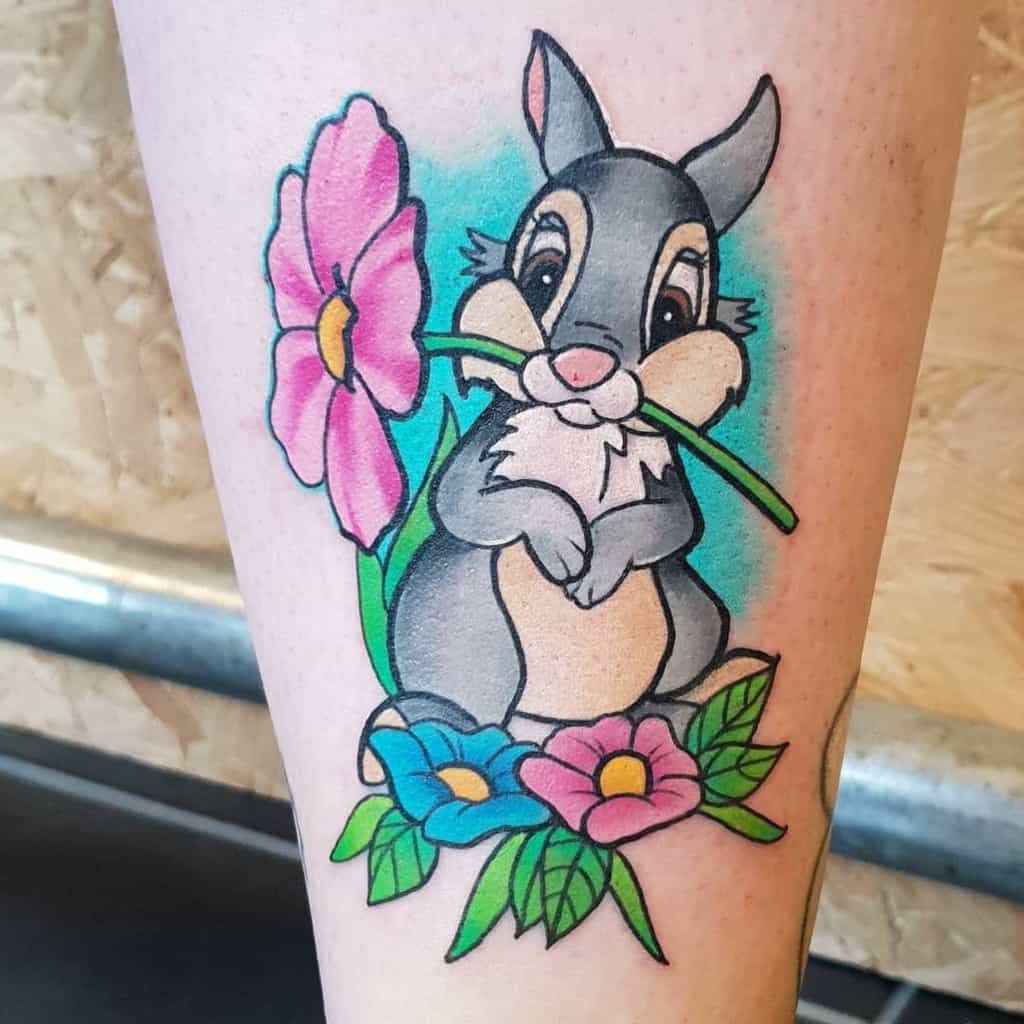 Tattoo of a cartoon rabbit holding a pink flower in its mouth, surrounded by two flowers and leaves, one pink and one blue