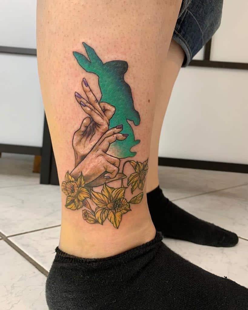 Tattoo featuring hands casting a blue green rabbit shadow, surrounded by yellow flowers