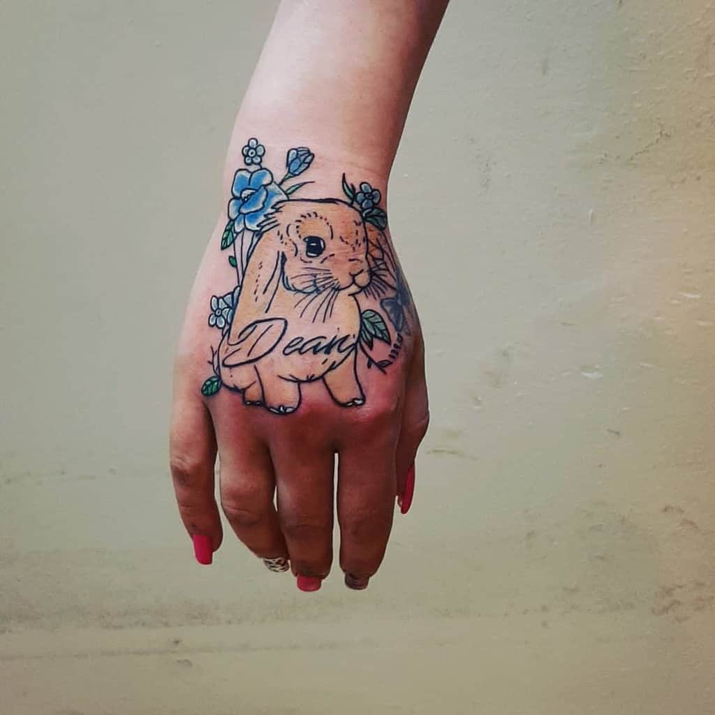 Charming tattoo of a rabbit surrounded by flowers and green leaves with the word 'Dear' written beneath it
