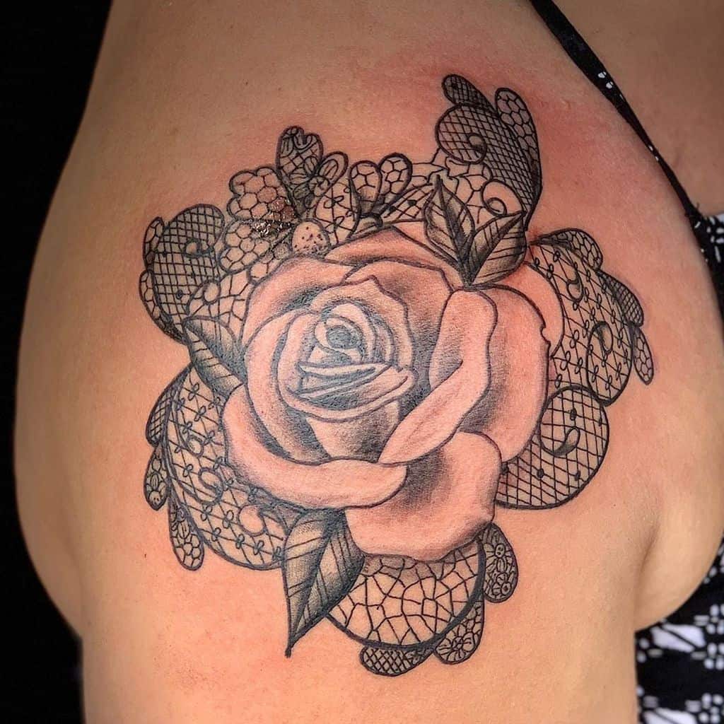 Black and grey shoulder tattoo featuring a shaded rose surrounded by intricate lace patterns, floral accents, and delicate netting.