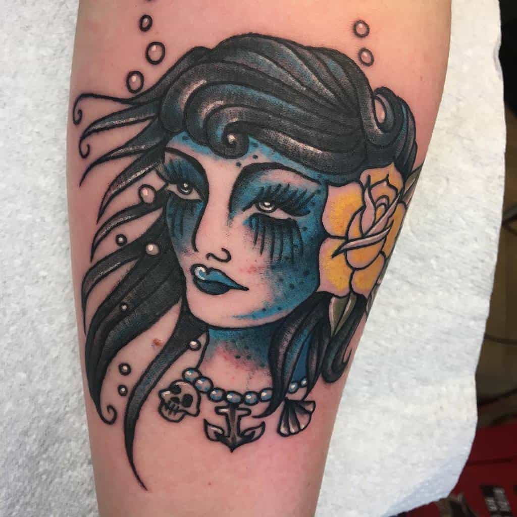 Neo-traditional tattoo of a blue-skinned siren with dark flowing hair, a yellow rose, and nautical-themed jewelry, evoking a mystical underwater vibe
