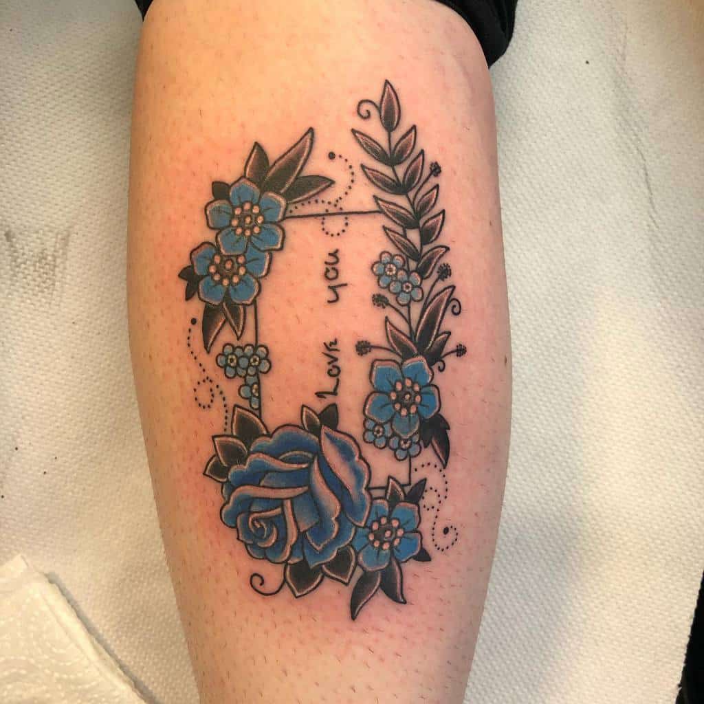 Tattoo of a blue rose and floral wreath on a forearm with the words "Love you" inside in a paper and dots