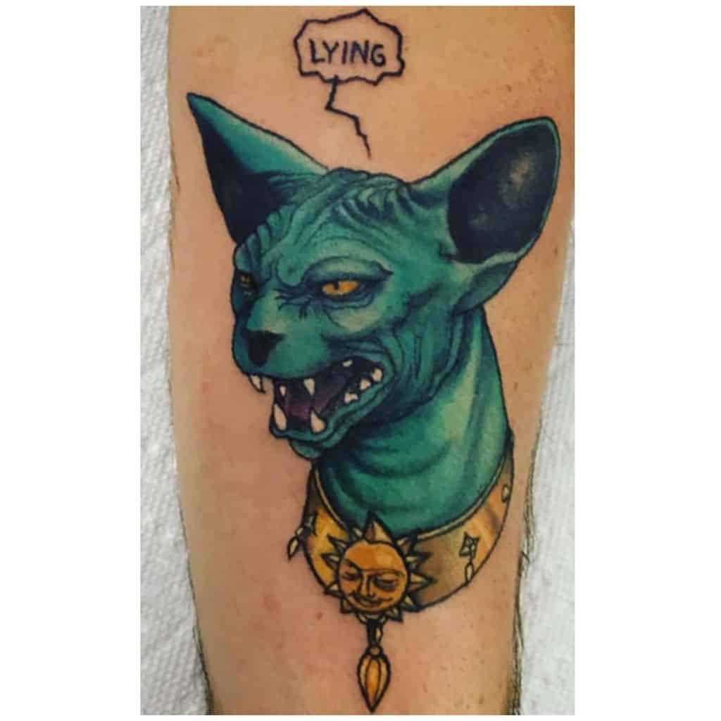 A colorful tattoo of a snarling blue Sphynx cat wearing a golden sun pendant, with a speech bubble saying 'LYING,' giving it a bold, mystical look.