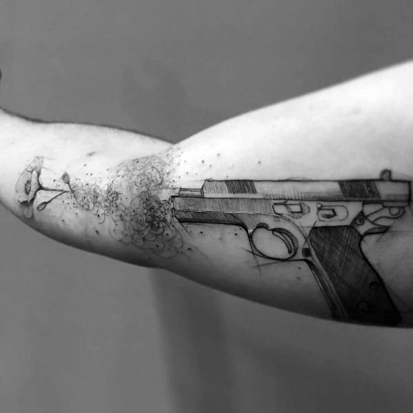 Black and gray forearm tattoo of a 1911 pistol, designed with a creative dispersion effect that gives the illusion of the gun disintegrating.