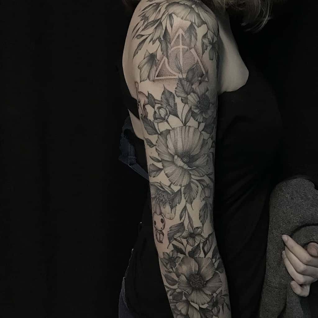 Black and grey full-sleeve floral tattoo with detailed flowers and leaves, featuring a subtle Deathly Hallows symbol on the shoulder.
