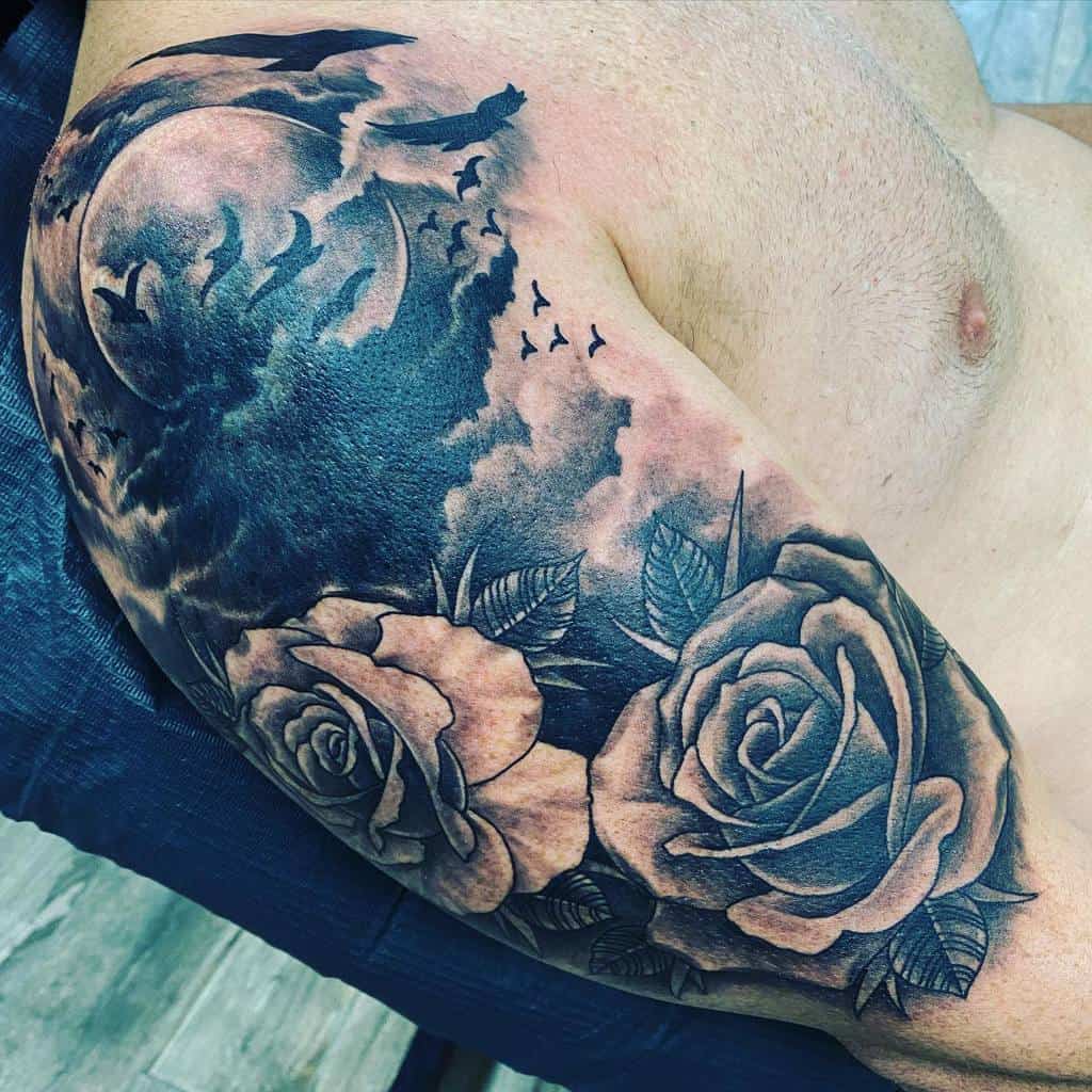 Arm tattoo featuring two roses, a moon, clouds, and flying birds in black and gray shading