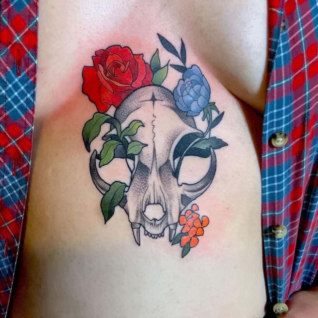 Tattoo of a cat skull with red, blue, and orange flowers on a person's chest