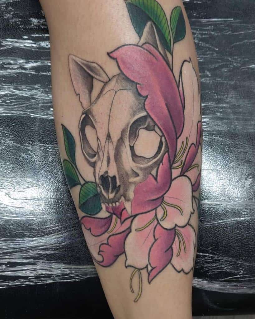A tattoo featuring a cat skull, intricately surrounded by pink flowers and green leaves