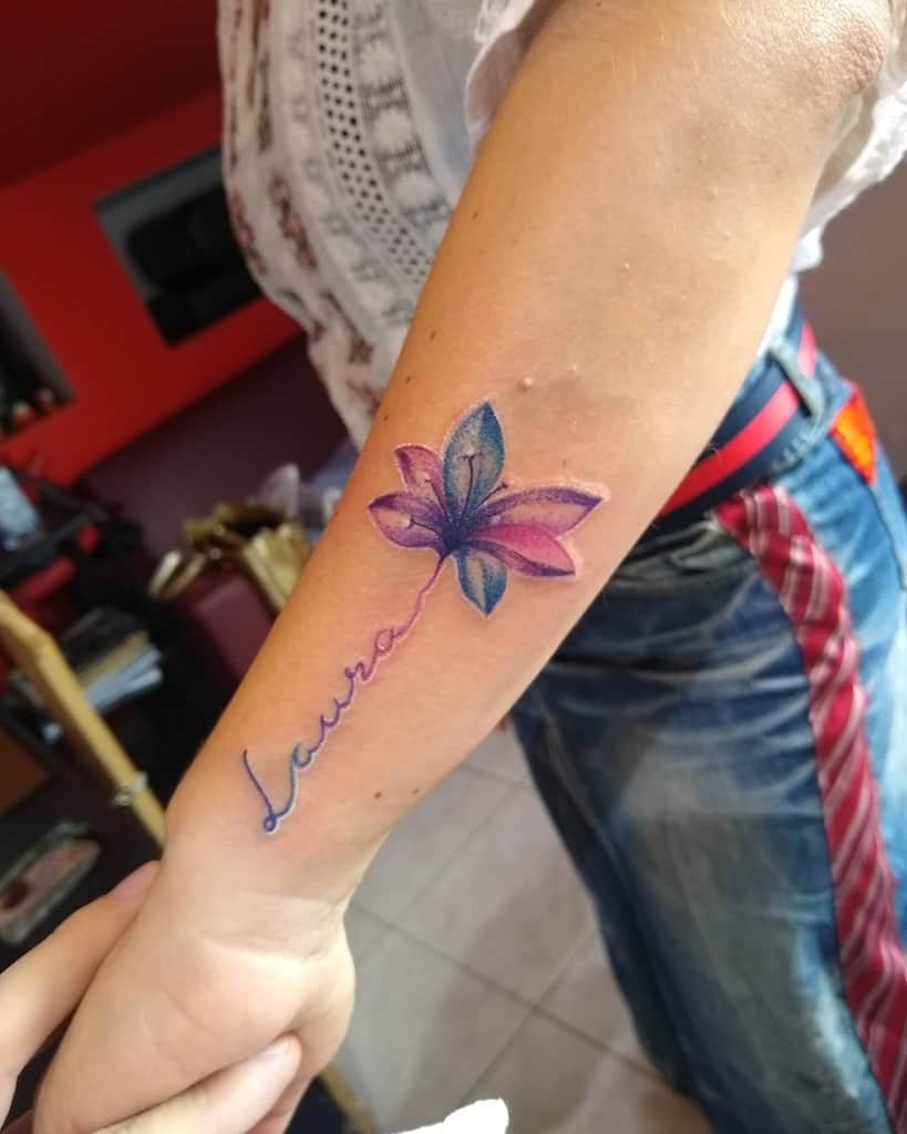 Tattoo of a pink and blue flower with the name "Laura" written in cursive, extending from the stem