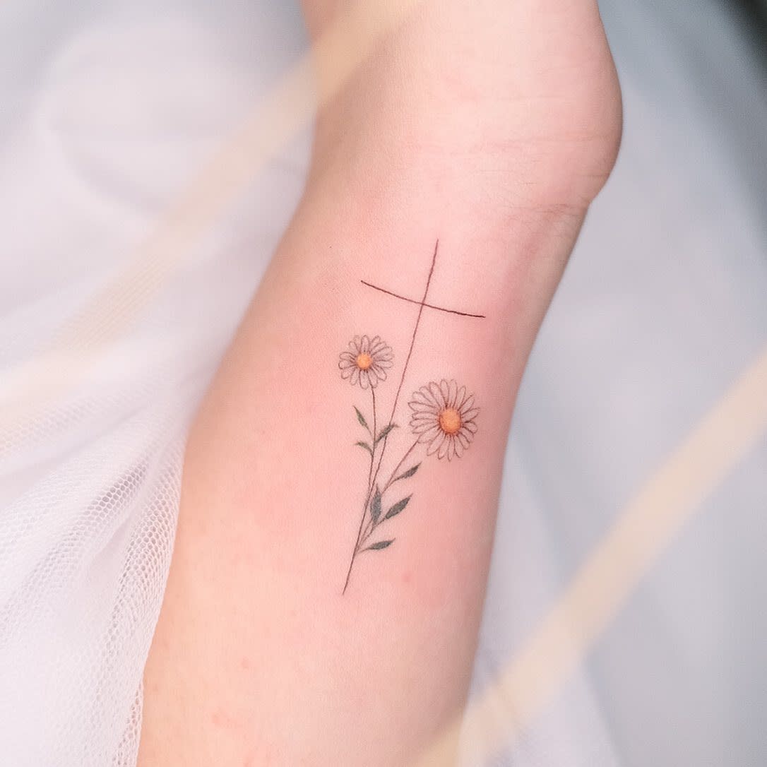 A delicate tattoo of two daisies intertwined with a subtle cross graces the forearm