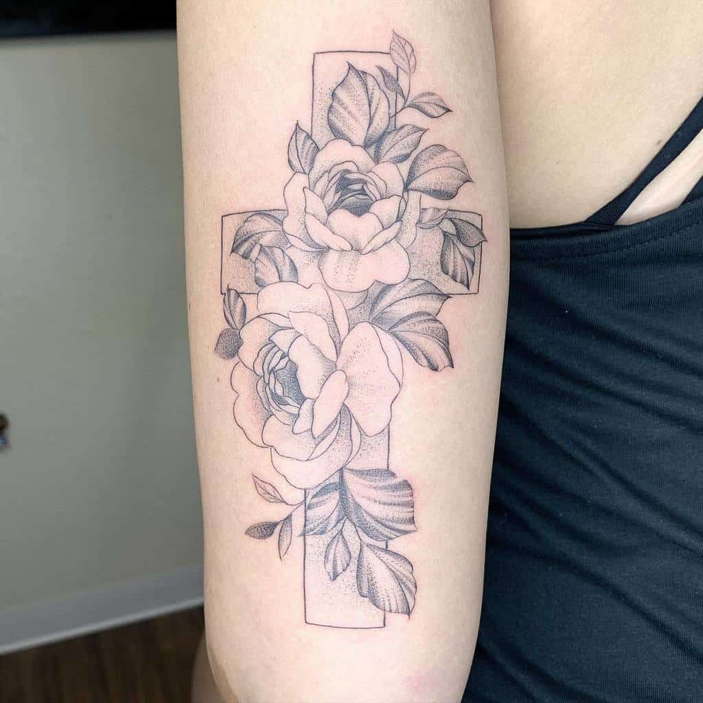 A stunning cross tattoo adorned with blooming roses and delicate leaves graces the upper arm, rendered beautifully in black and gray ink