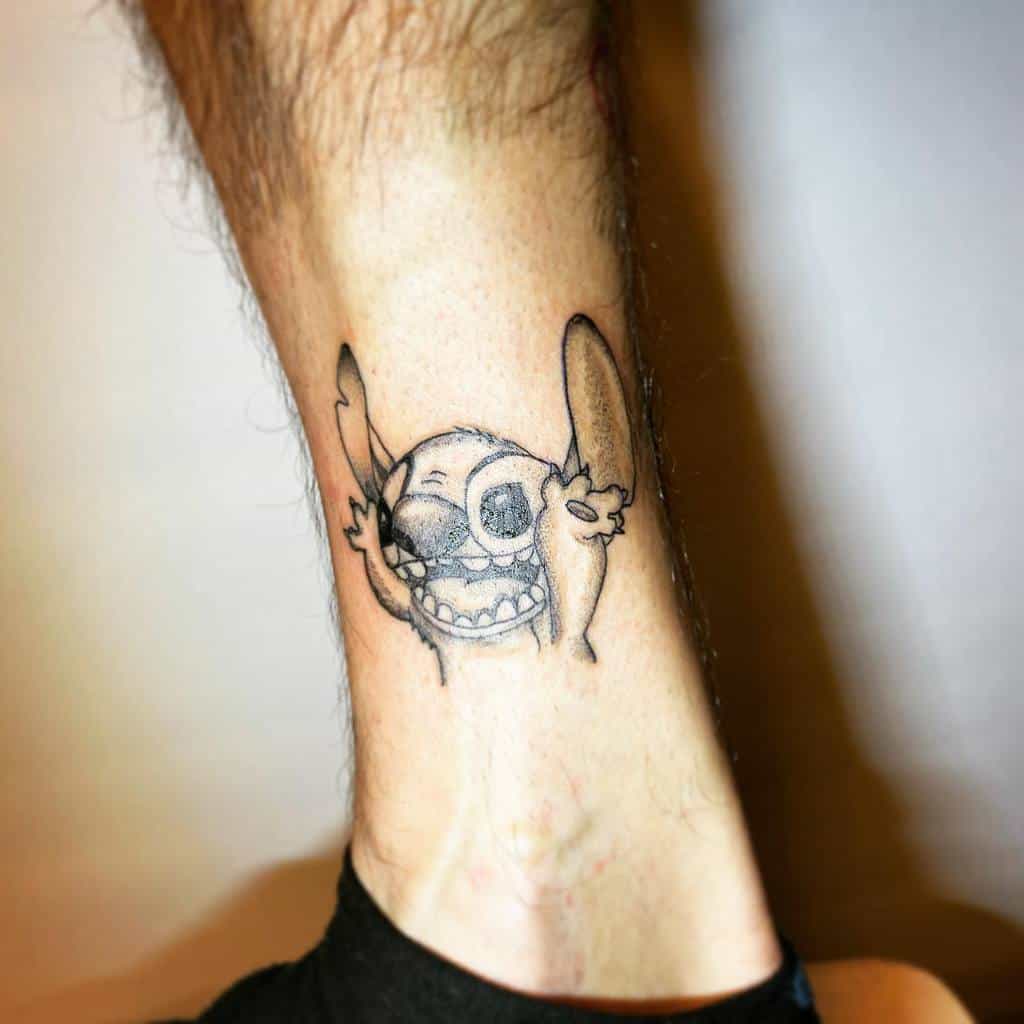 Black and white tattoo of Stitch from Lilo & Stitch with a playful expression, raising his arms in excitement, on the ankle.