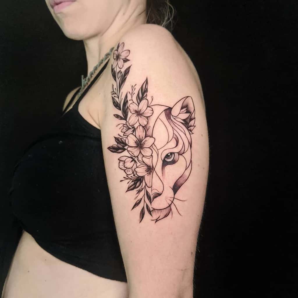 Black and grey lioness tattoo on the upper arm, featuring a half-face design with bold geometric lines and delicate floral details for contrast.