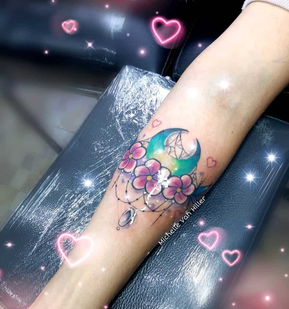 Tattoo of a crescent moon with pink flowers and a hanging gem on forearm
