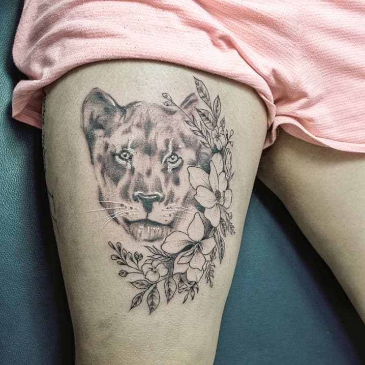 Black and grey lioness thigh tattoo with soft shading, floral details, and fine linework, blending strength and femininity in an elegant design.