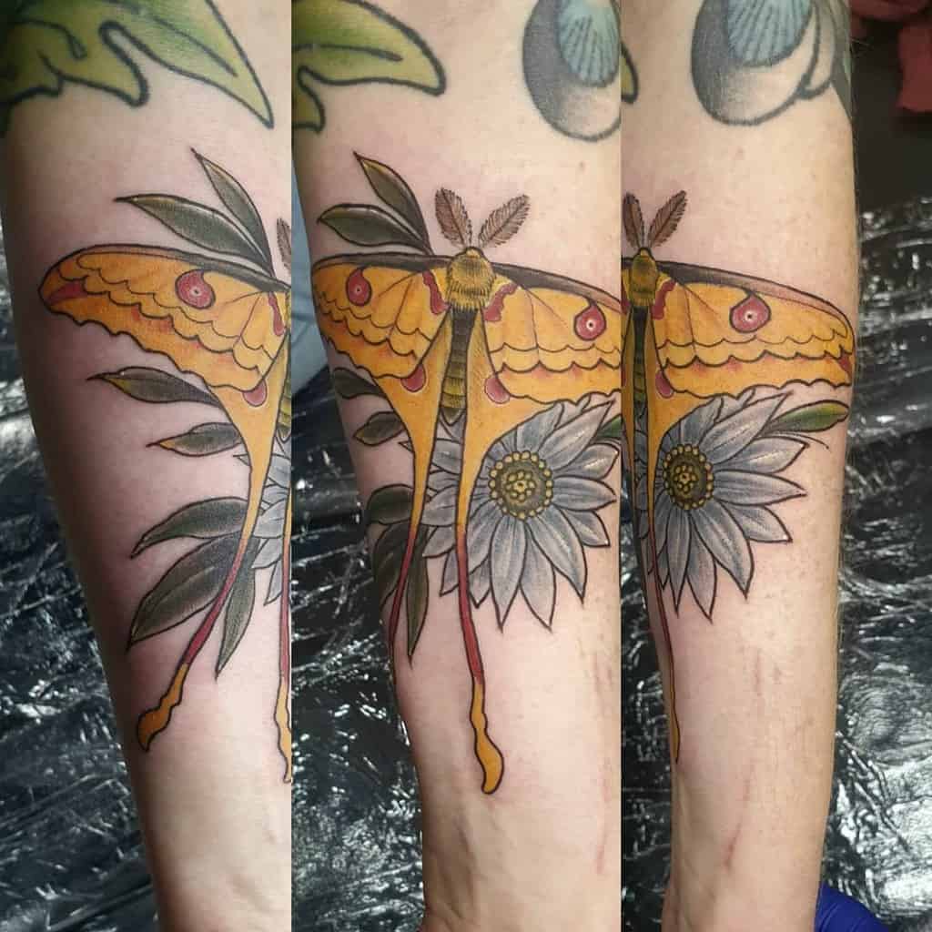 A colorful Luna Moth tattoo with yellow and orange wings, surrounded by leaves and daisy flowers in grey tones
