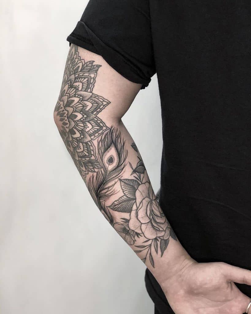 Black and grey forearm tattoo featuring a peacock feather, floral elements, and intricate mandala patterns with fine linework and shading.