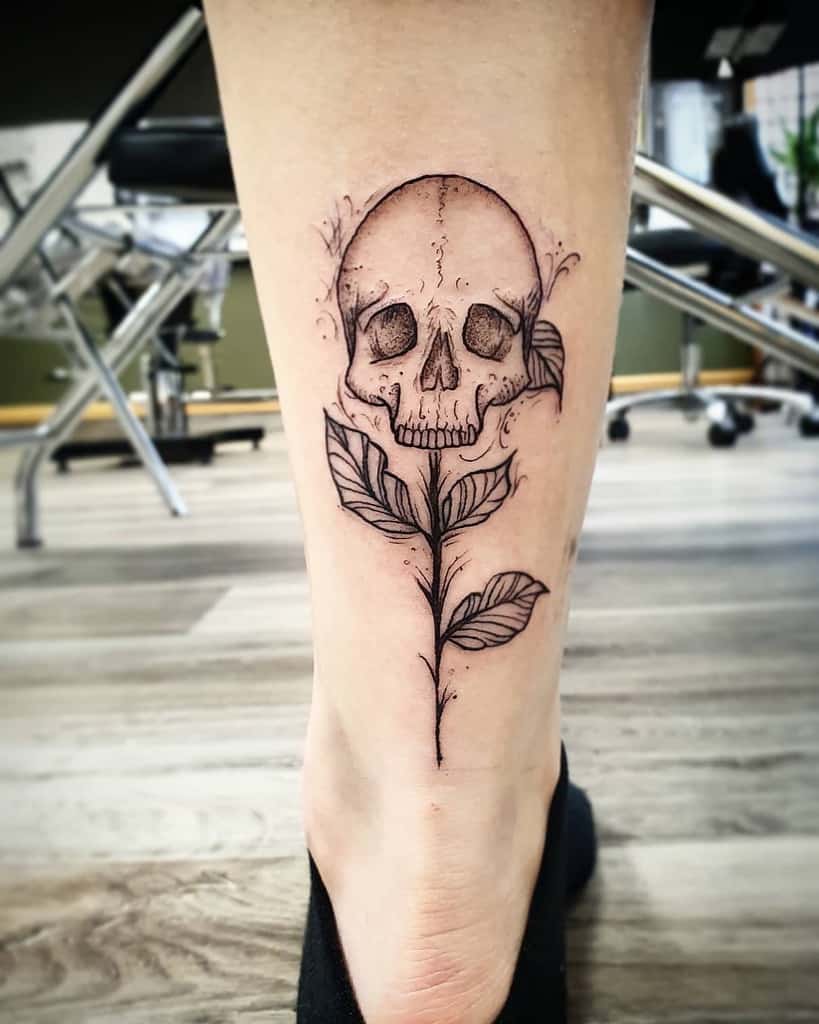 Black and grey tattoo of a skull growing from a delicate stem with leaves, blending nature and mortality, inked on the lower leg.
