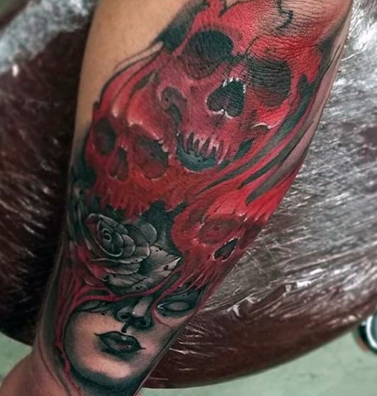 Forearm tattoo featuring red and black skulls, flames, a rose, and a woman's face