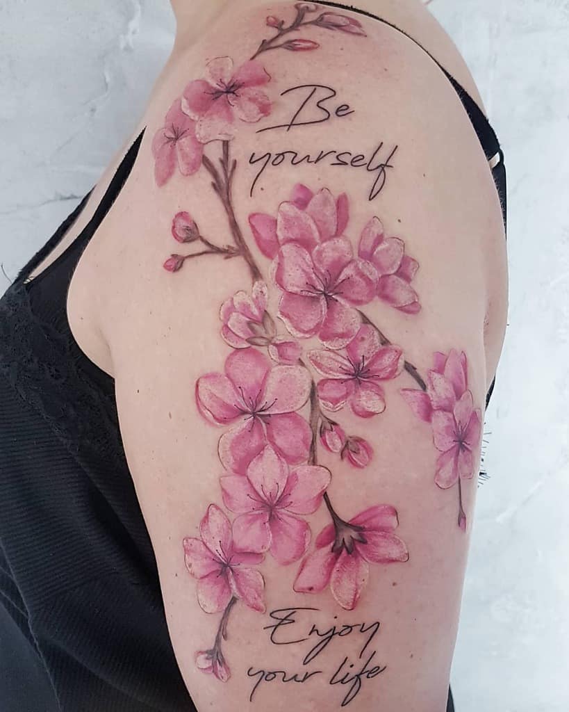 Tattoo of pink cherry blossoms on an upper arm, with cursive text saying "Be yourself" and "Enjoy your life"