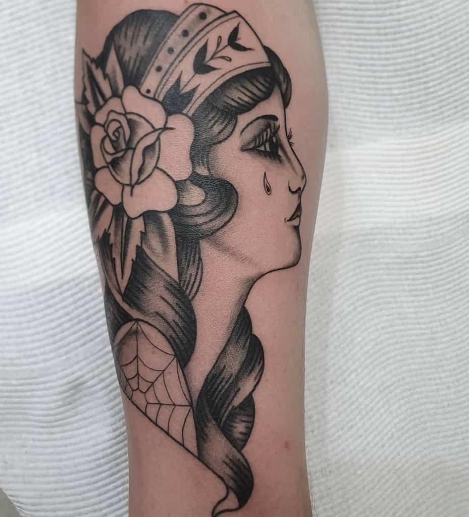 Black and grey tattoo of a gypsy woman with a rose, a tear drop, and braided hair, featuring bold shading and classic details on the forearm.