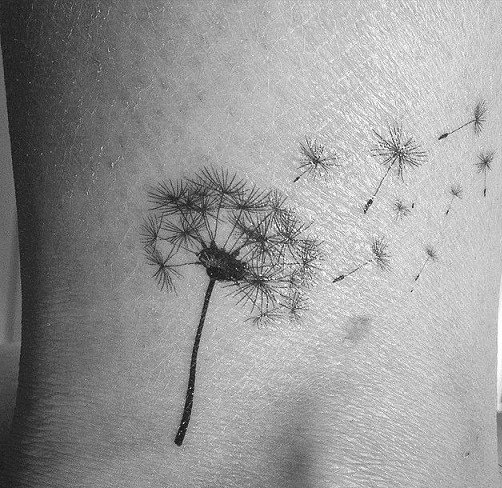 This tattoo design showcases a dandelion with its seeds floating away, captured in delicate black ink. The floating seeds symbolize the idea of release and freedom, combining natural beauty with a sense of lightness and movement.