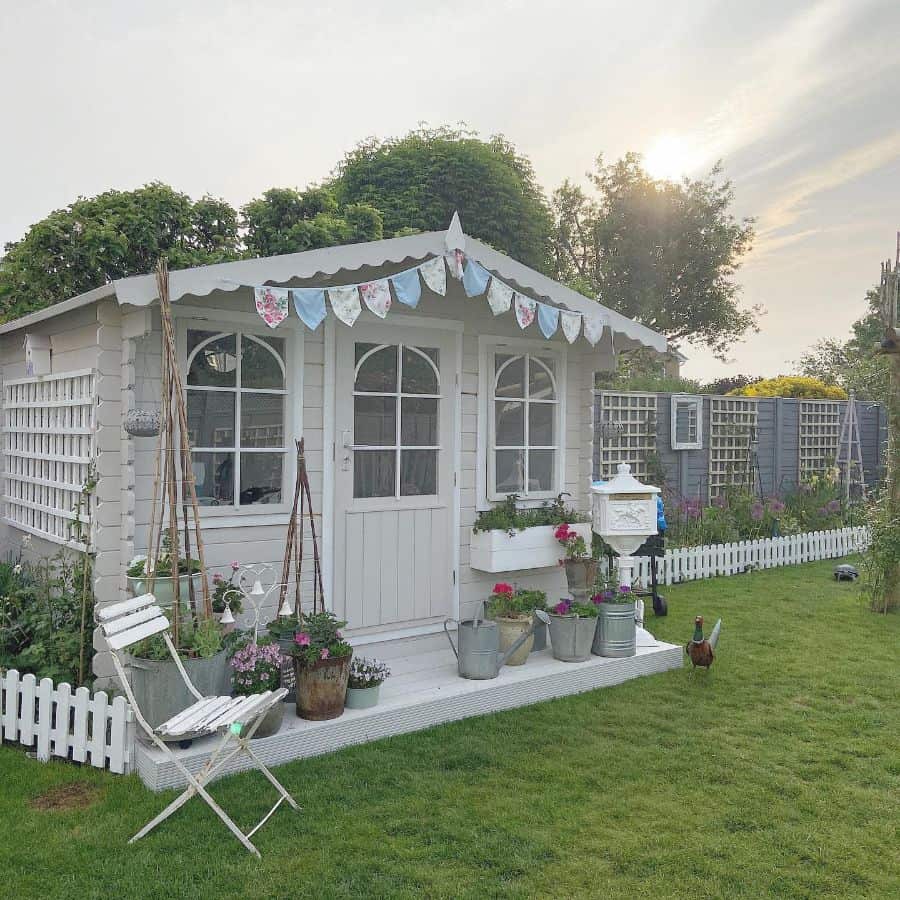 Small garden shed with a white picket fence, surrounded by plants, sunlight filters through trees nearby