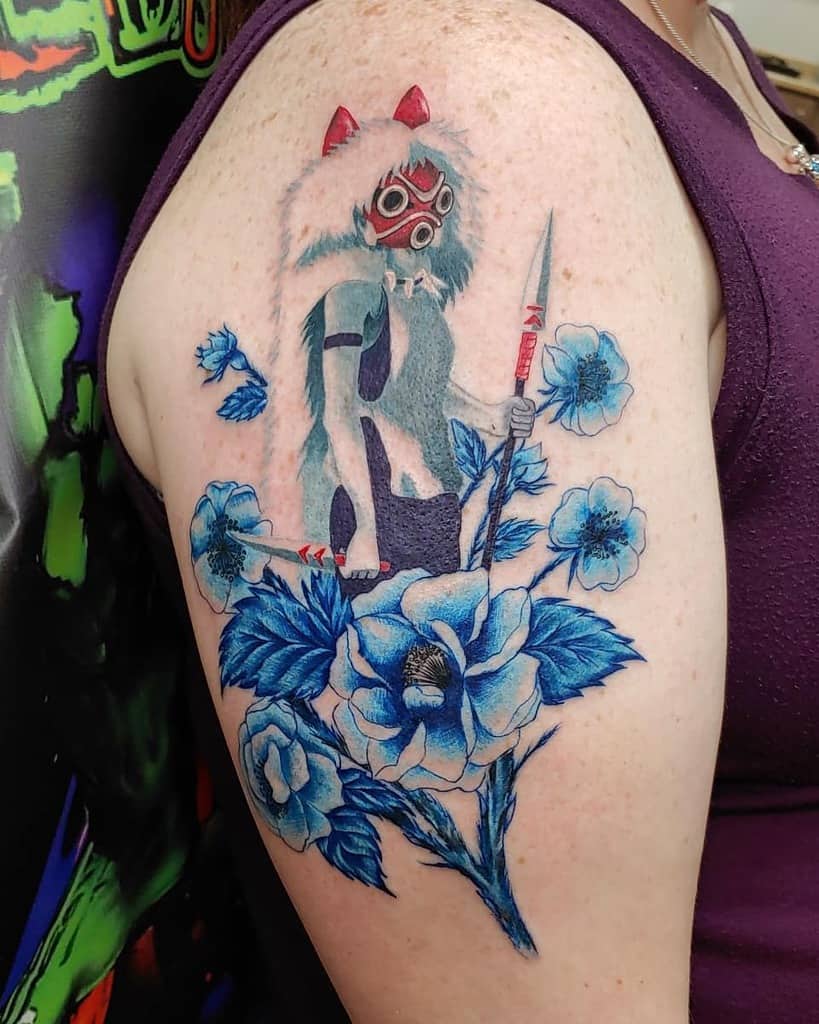 Vibrant tattoo of San in her wolf mask, holding a spear, surrounded by large blue flowers and leaves, with dynamic shading and soft details.