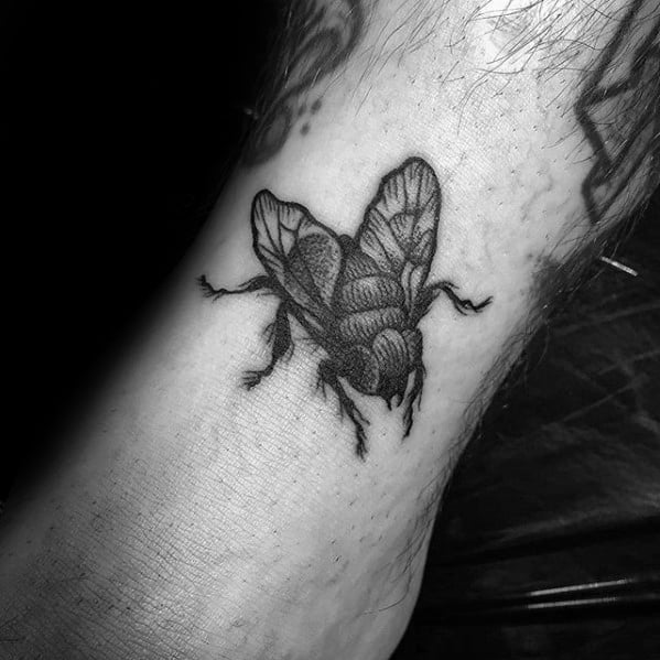 Realistic black and grey fly tattoo on the ankle with detailed shading and textured wings, creating a lifelike appearance.