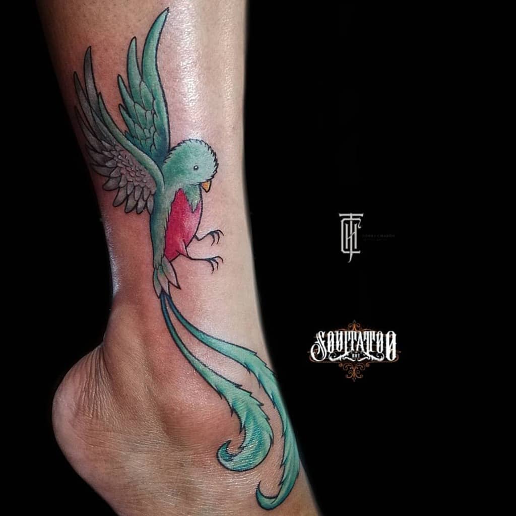 Elegant quetzal tattoo on the ankle, featuring a stylized green and red bird in flight with long, flowing tail feathers and soft shading