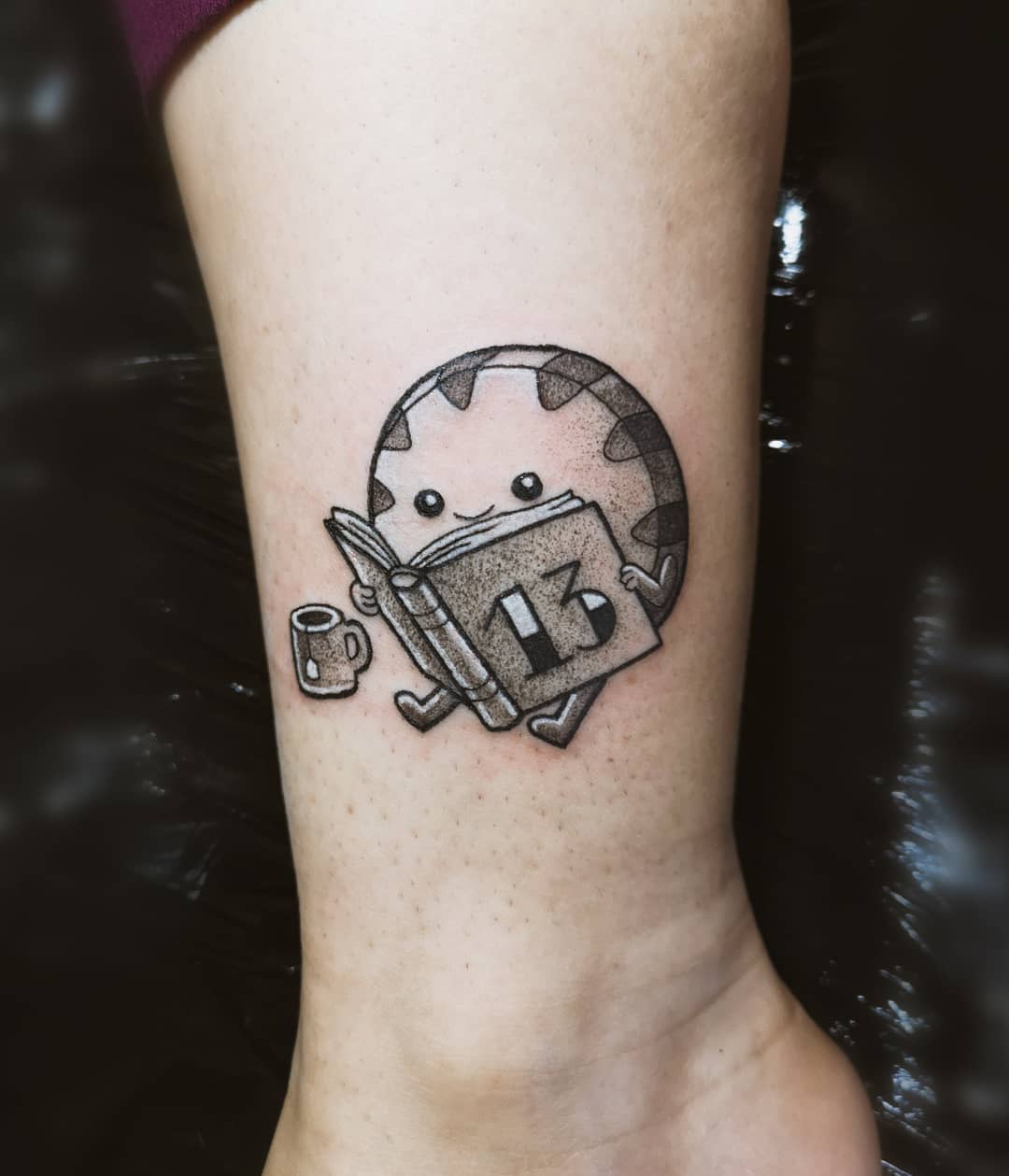 Tattoo of a cute candy character reading a book labeled "13, with a small cup and teabag beside it