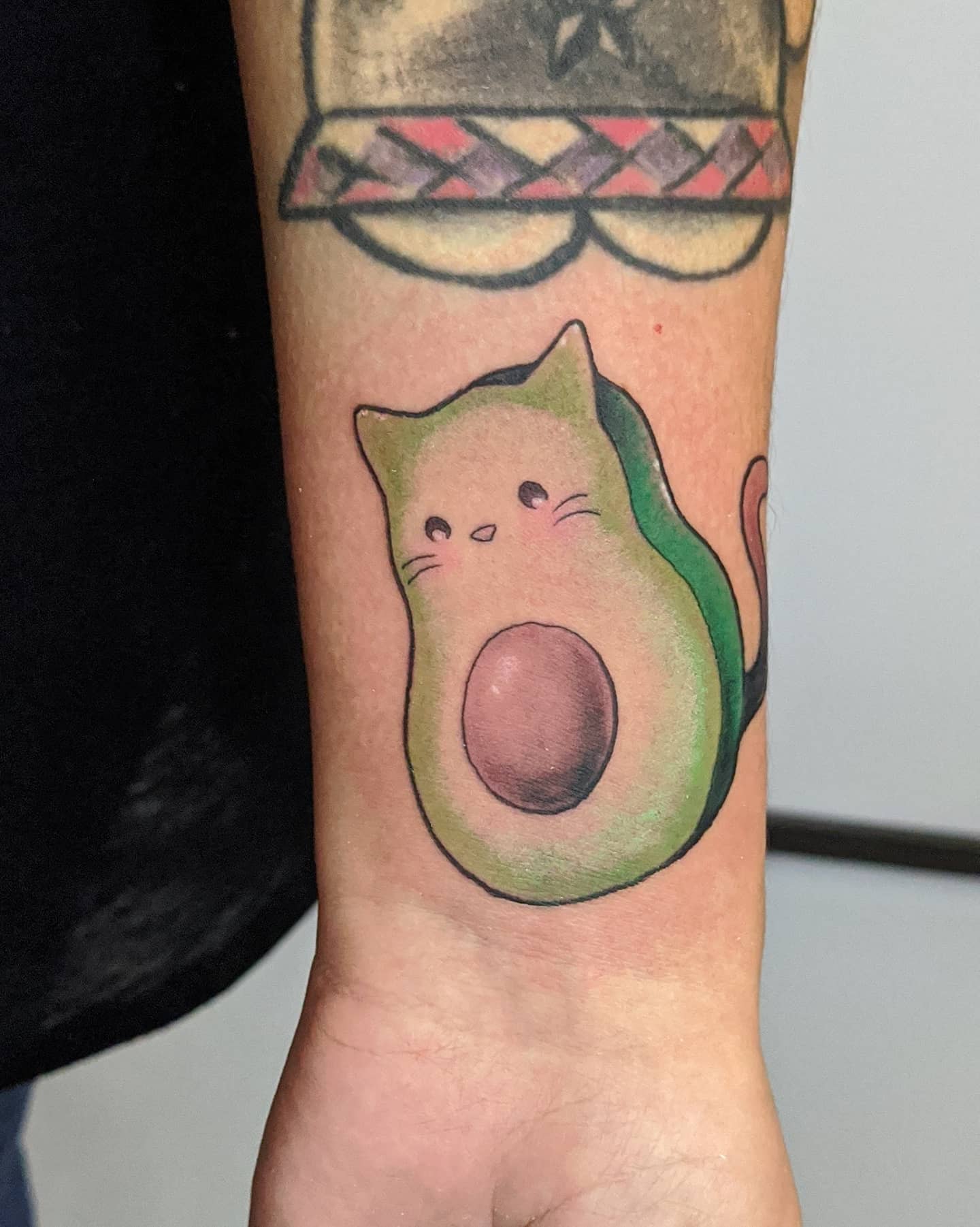 Tattoo of an avocado with a cute cat face and ears, under a partially visible geometric tattoo