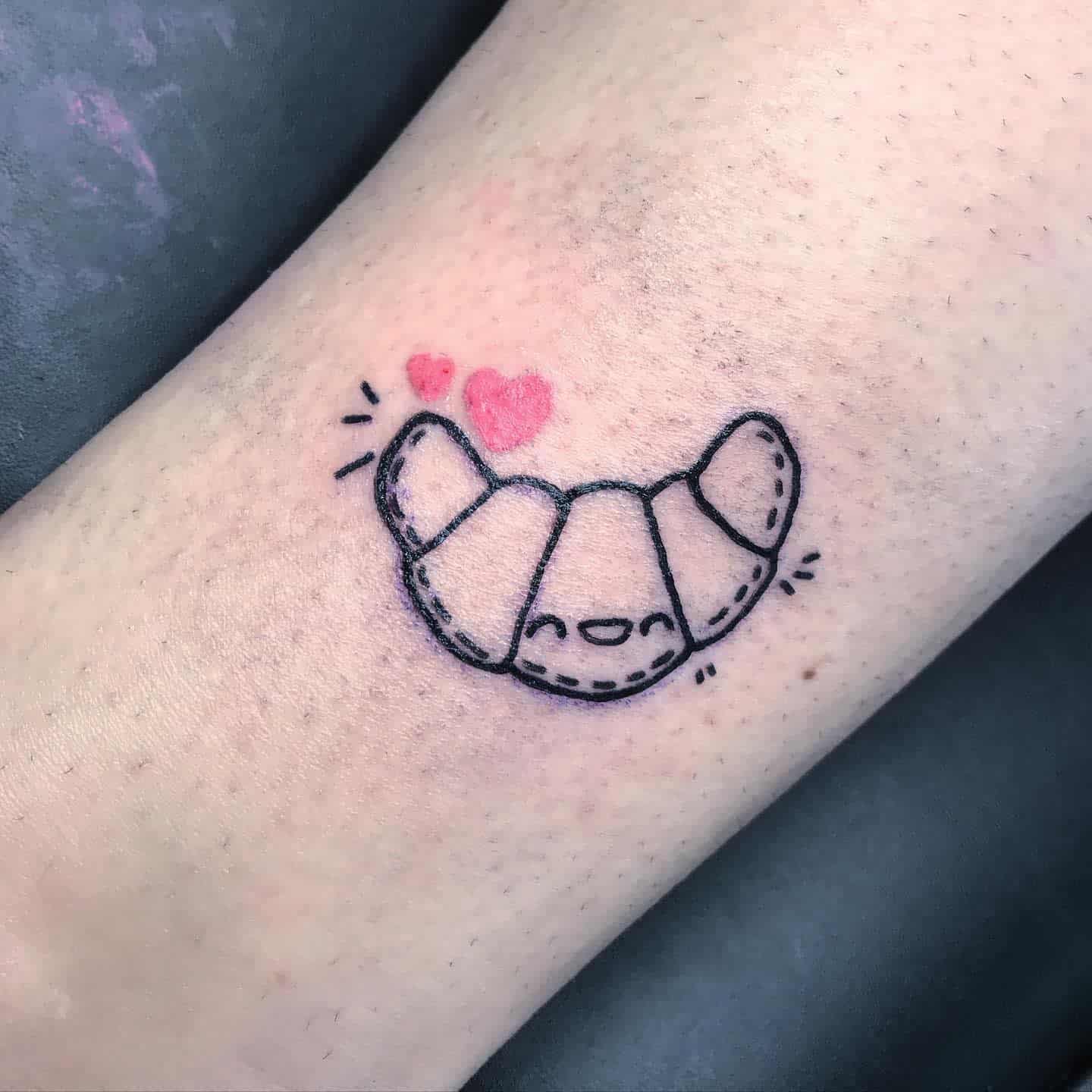 Tattoo of a smiling croissant with pink hearts above it