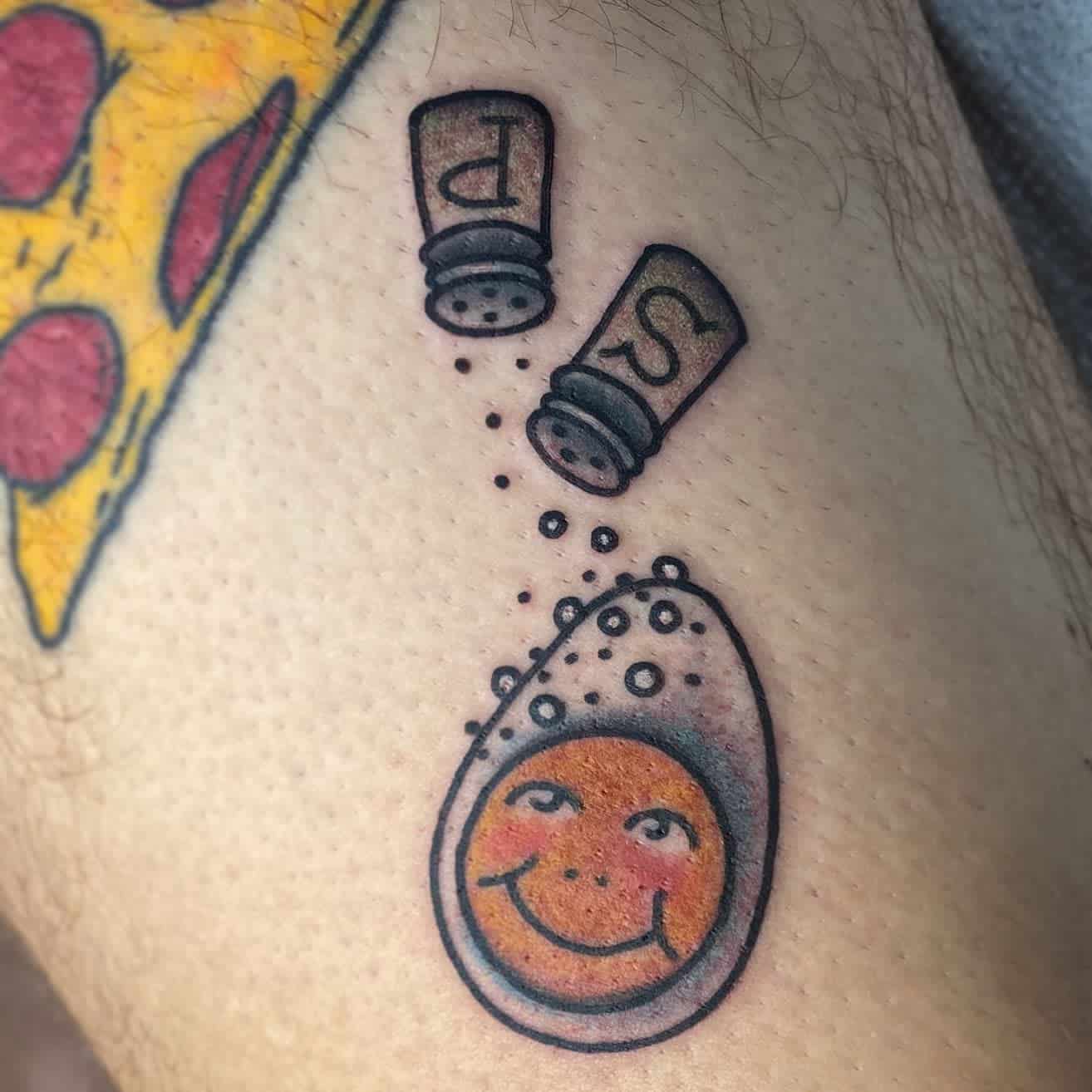 Tattoo of a cracked egg with a smiling yolk, sprinkled with salt and pepper, next to a pizza slice tattoo