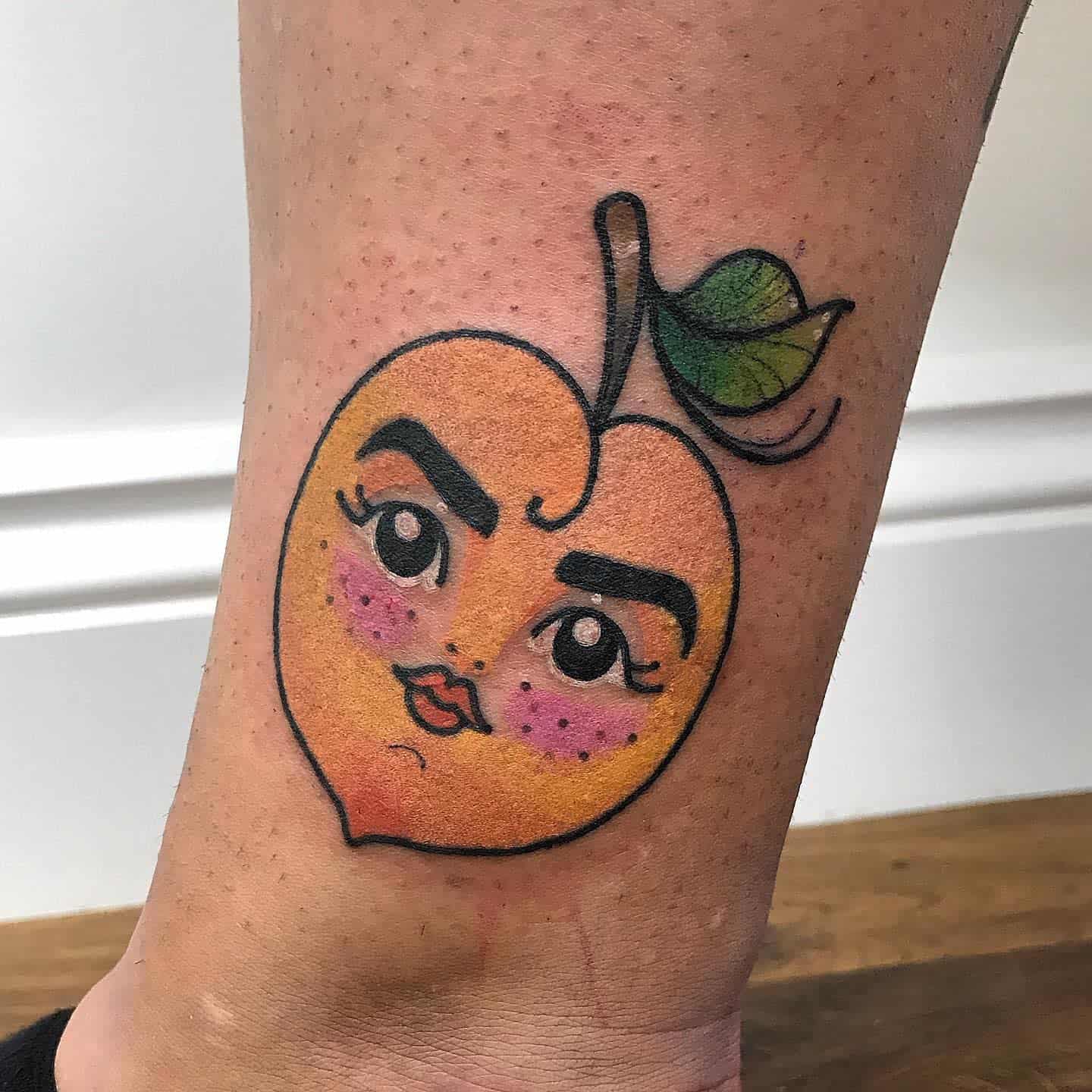 Tattoo of a peach with a face, bold eyebrows, and red cheeks, featuring a leaf and a stem