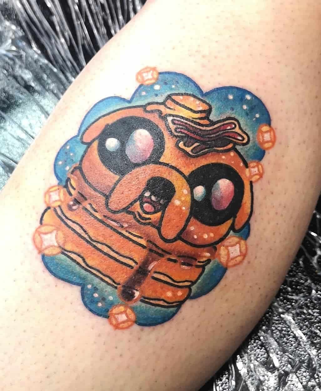 Tattoo of a cartoon-style dog resembling pancakes with syrup, butter, and bacon, surrounded by cubes and blue cloud background