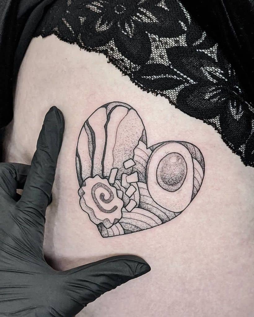 Black and white tattoo of a ramen bowl in a heart shape, featuring Naruto fishcake, boiled eggs, noodles, and green onions