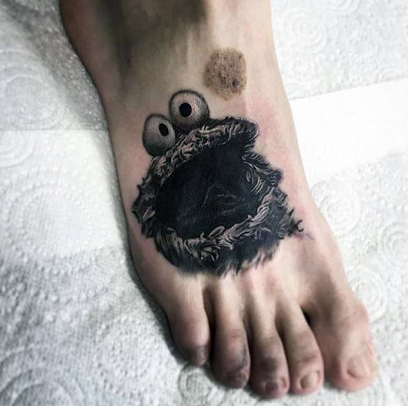 Black and grey Cookie Monster foot tattoo with wide eyes and open mouth, reaching for a realistic cookie above his head.