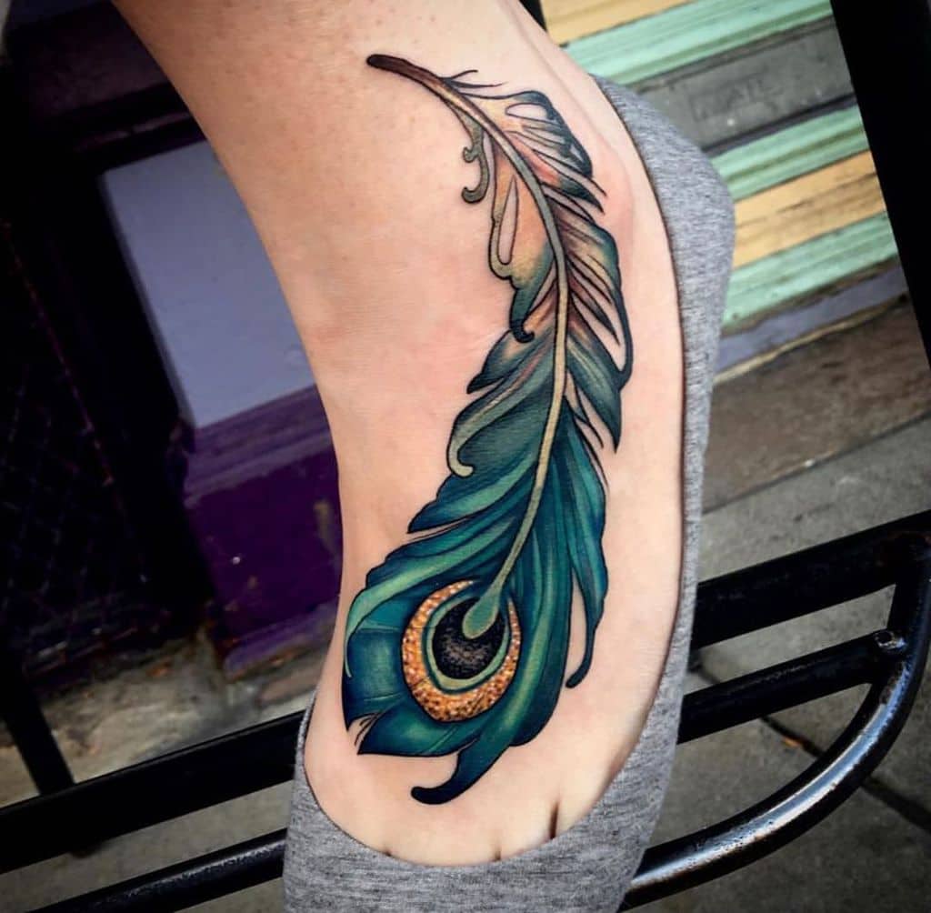 Vibrant peacock feather tattoo on the foot, featuring rich blue, green, and gold tones with intricate shading and fine detailing.