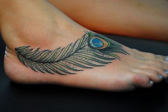Elegant peacock feather tattoo on the foot, featuring fine black outlines, subtle shading, and vibrant blue, green, and gold hues in the eye pattern.