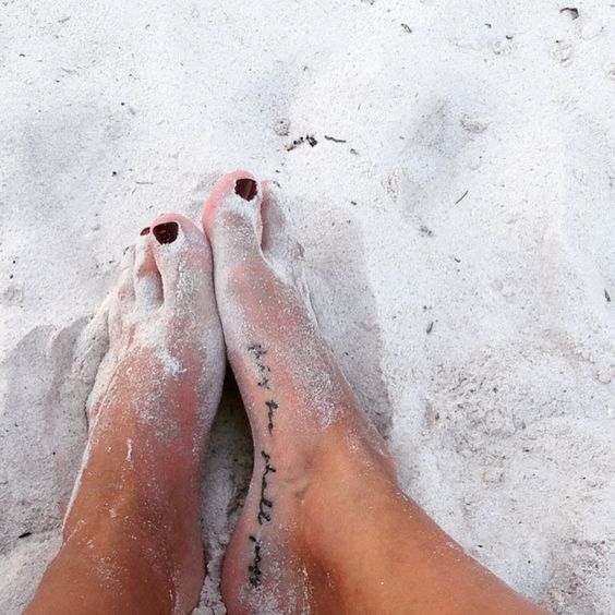 A "This Too Shall Pass" tattoo in cursive on a person's foot, with sand around the feet.