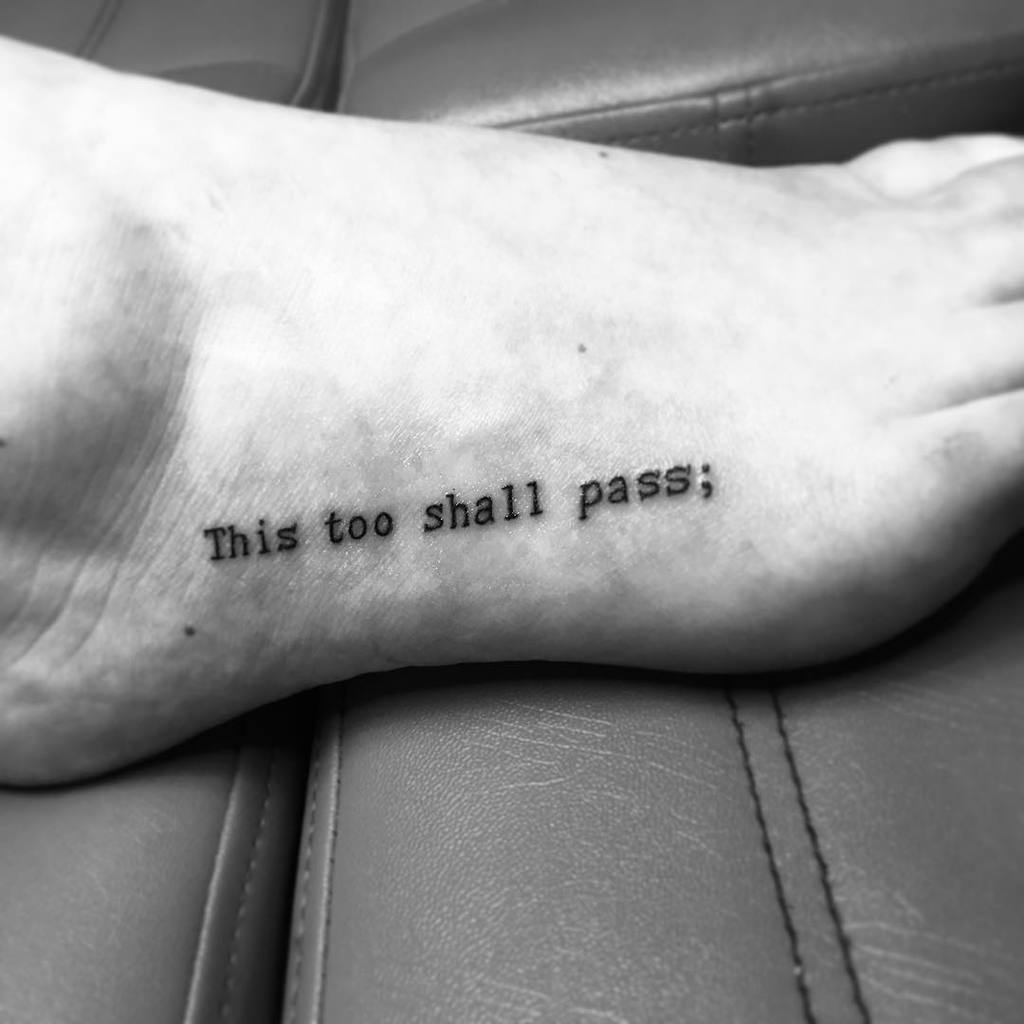 This image shows a tattoo on the foot with the phrase "This too shall pass;" in bold, typewriter-style font.