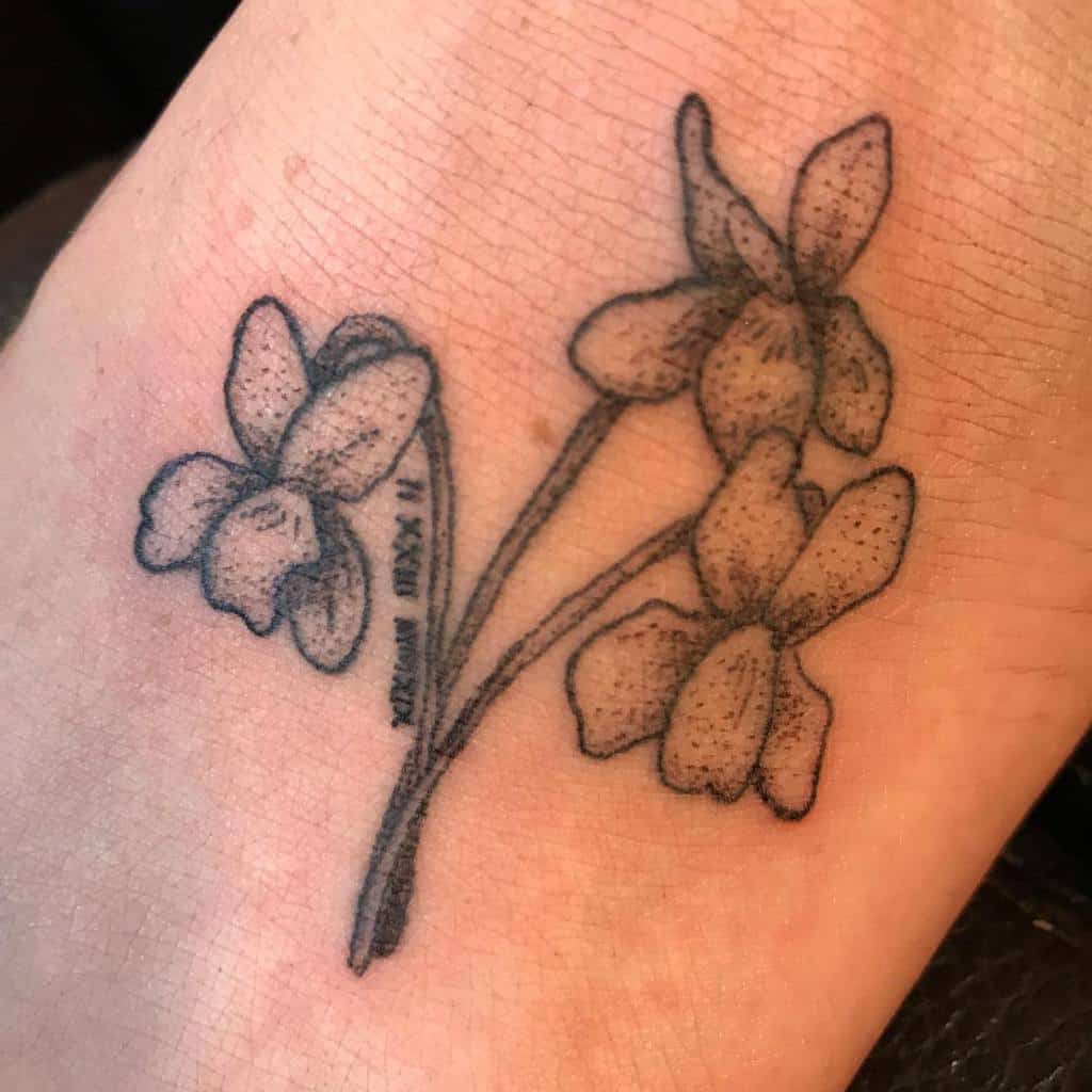 Minimalist black dotwork tattoo of delicate violet flowers, featuring fine details and a small inscription along the stem