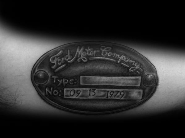 Realistic black and grey Ford Motor Company plaque tattoo on forearm, featuring engraved text, shading, and a vintage metal plate design.