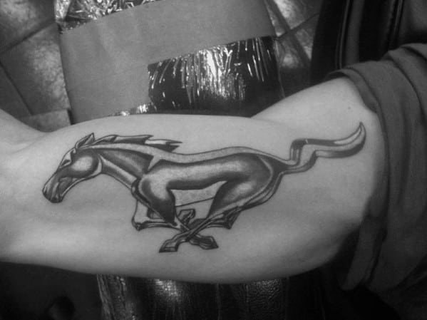 Bold black and grey Ford Mustang logo tattoo on inner arm, featuring the iconic running horse with dynamic shading and strong outlines.