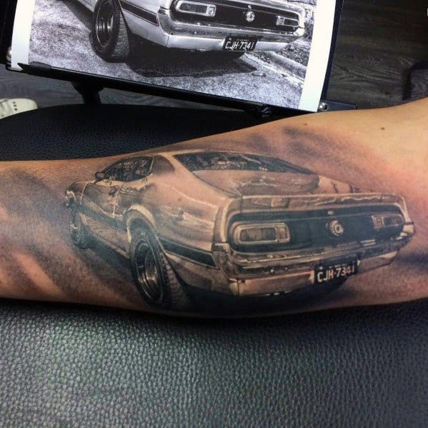 Realistic black and grey Ford Mustang tattoo on forearm, showcasing a detailed rear view with intricate shading and a vintage muscle car design.